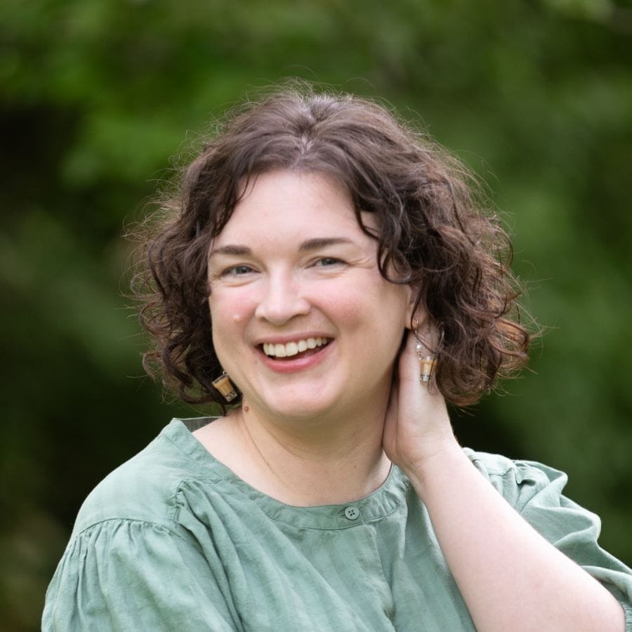 A woman with curly brown hair wearing a light green blouse smiles at the camera while touching her neck, with a blurred green background behind her.