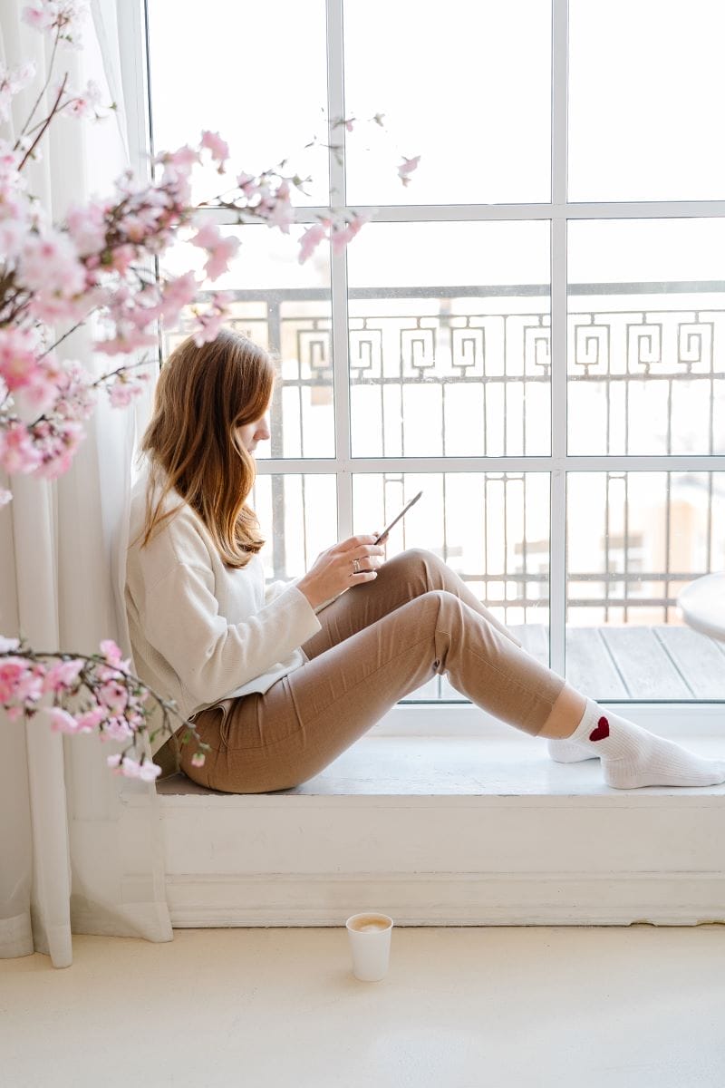 A woman sits on a windowsill, using her phone—perhaps researching an affiliate program or web design—as a cup of coffee rests on the floor and pink flowers bloom in the foreground.