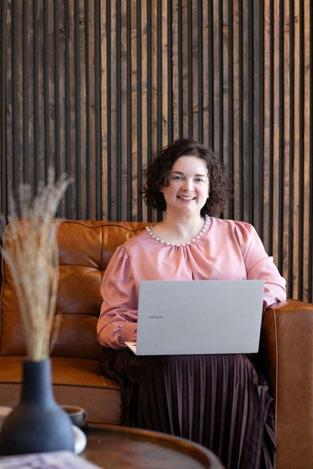 A woman with curly brown hair, wearing a pink blouse, sits on a brown leather sofa with a laptop on her lap. Wood paneling is visible in the background.