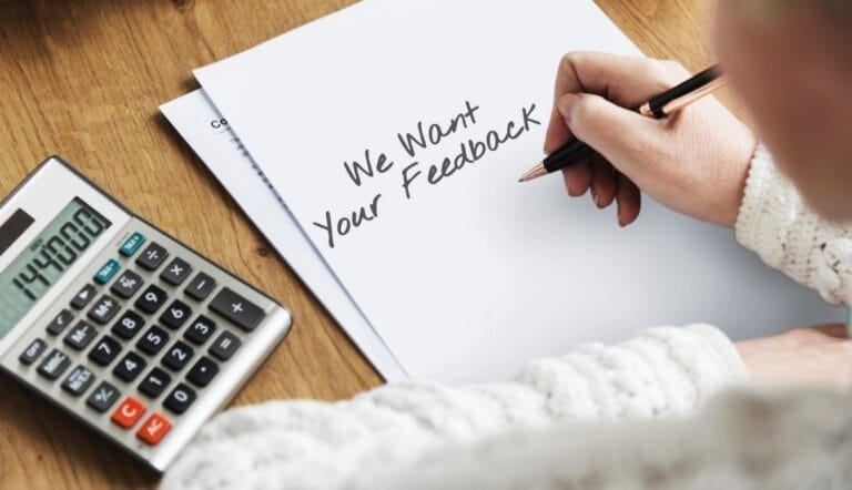 A person writes "We Want Your Feedback" on a sheet of paper next to a calculator and another document on a wooden table.
