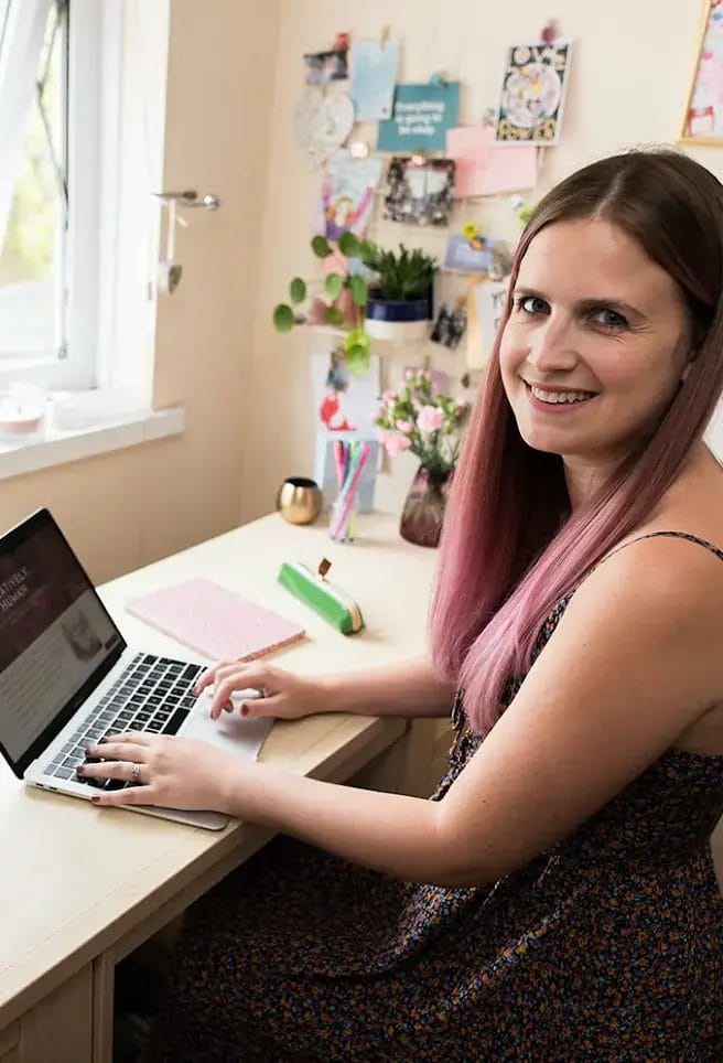 A woman with long, straight hair sits at a desk using a laptop, smiling at the camera. The desk has notebooks, flowers, and decor on the wall behind her.