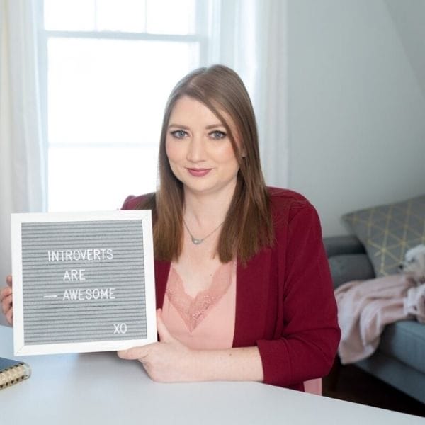 A woman sits at a desk holding a sign that reads "INTROVERTS ARE AWESOME XO" in a bright room with a couch and window in the background.