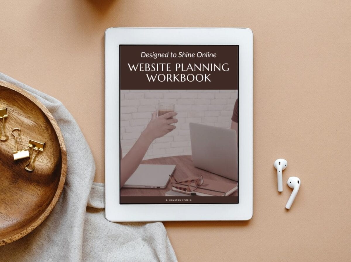 A tablet displays a “Website Planning Workbook” cover, showing a person holding a drink near a laptop; AirPods, a dish, and office supplies are nearby on a beige surface.