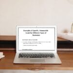 A laptop on a wooden desk displays a slide titled “Examples of Specific, Measurable Goals for Different Types of Businesses,” with two sample goals listed.