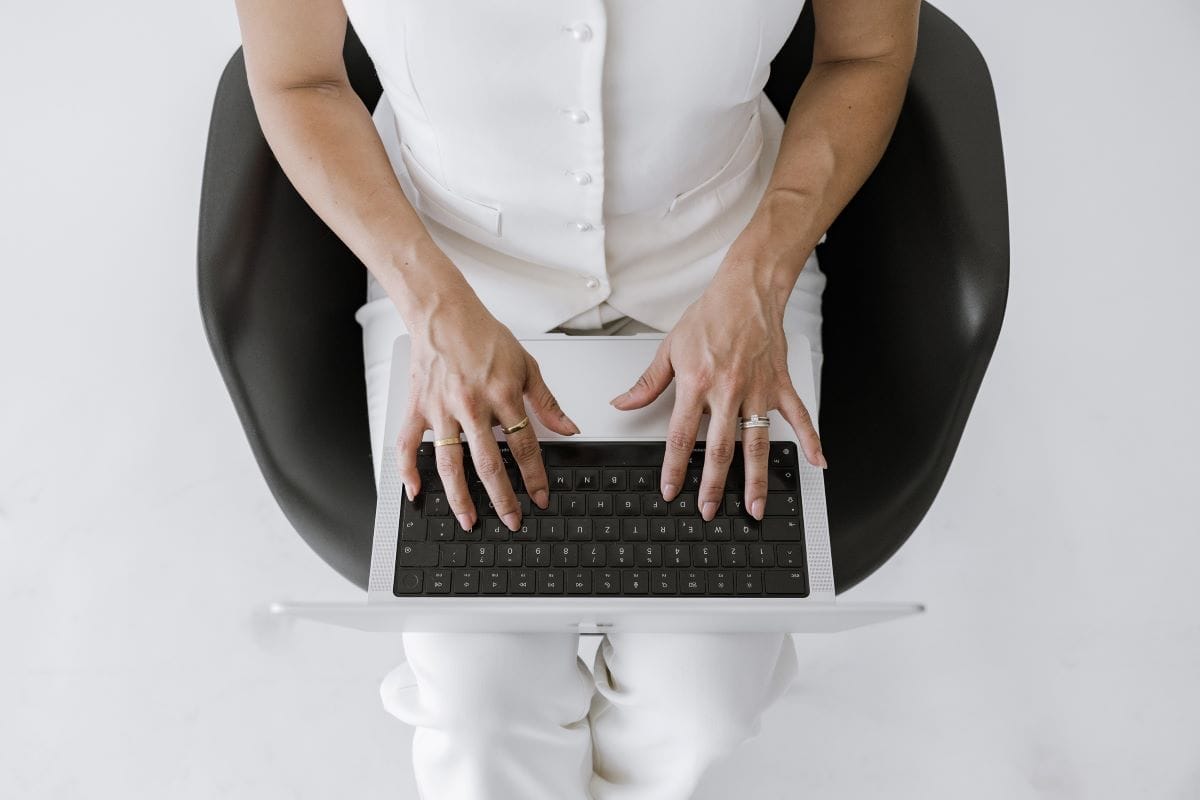 A person wearing white clothing sits in a black chair and types on a laptop, viewed from above.