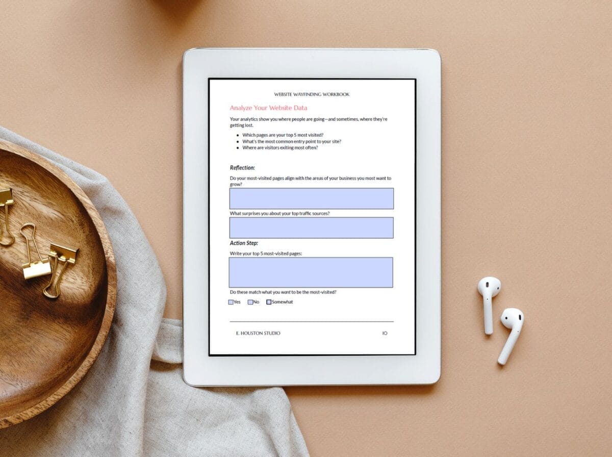 A tablet displaying a worksheet titled "Analyze Your Website Data" on a beige surface, next to a wooden tray with clips, a cloth napkin, and a pair of wireless earbuds.