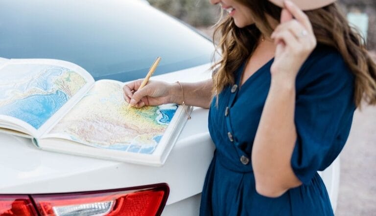 Woman in a blue dress stands by a white car, holding a hat and marking a location on a map book with a pencil—just like following website navigation best practices for finding your way.