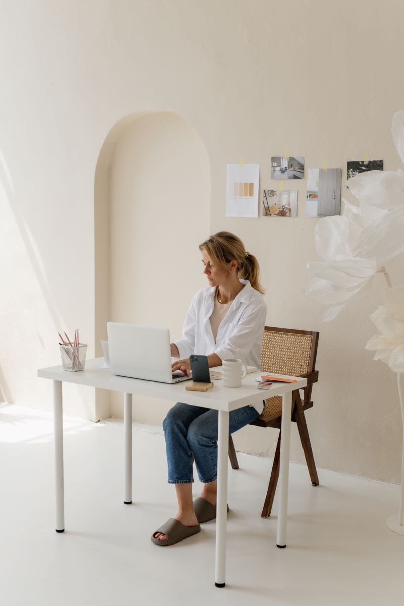 A woman sits at a white desk using a laptop in a minimal, light-colored room with papers and photos on the wall, focused on website implementation services.