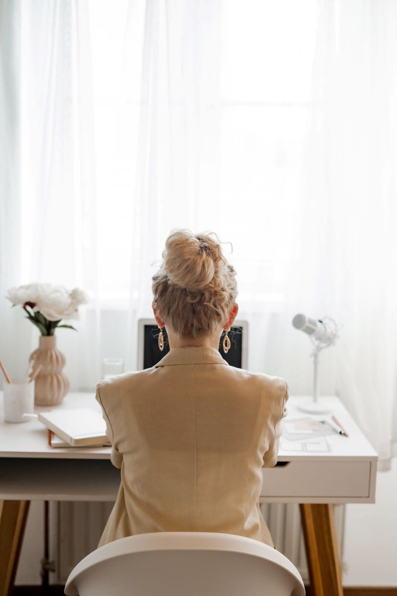 A person with blonde hair in a bun sits at a white desk facing a laptop, ready to provide website implementation services, with a vase of flowers, microphone, and notebooks nearby in a bright room.