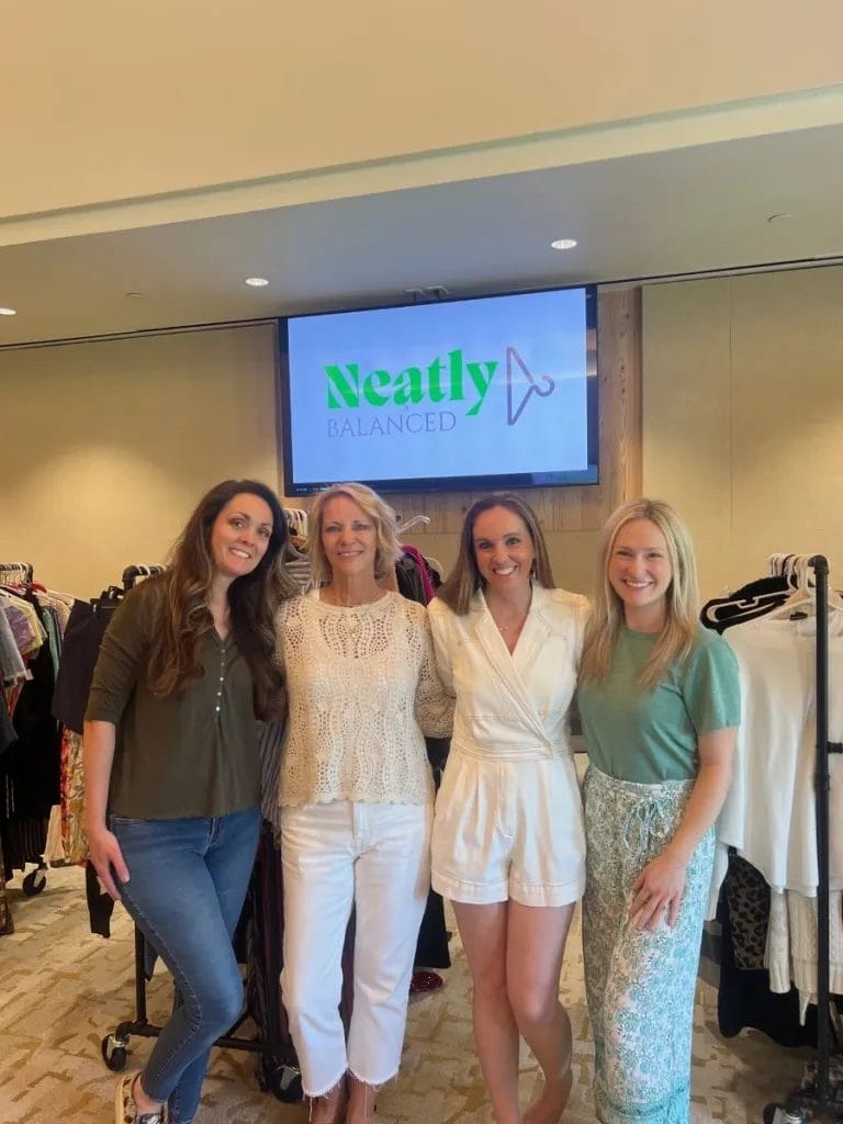 Four women stand smiling in front of a clothing rack and a screen displaying the "Neatly Balanced" logo in a well-lit room.