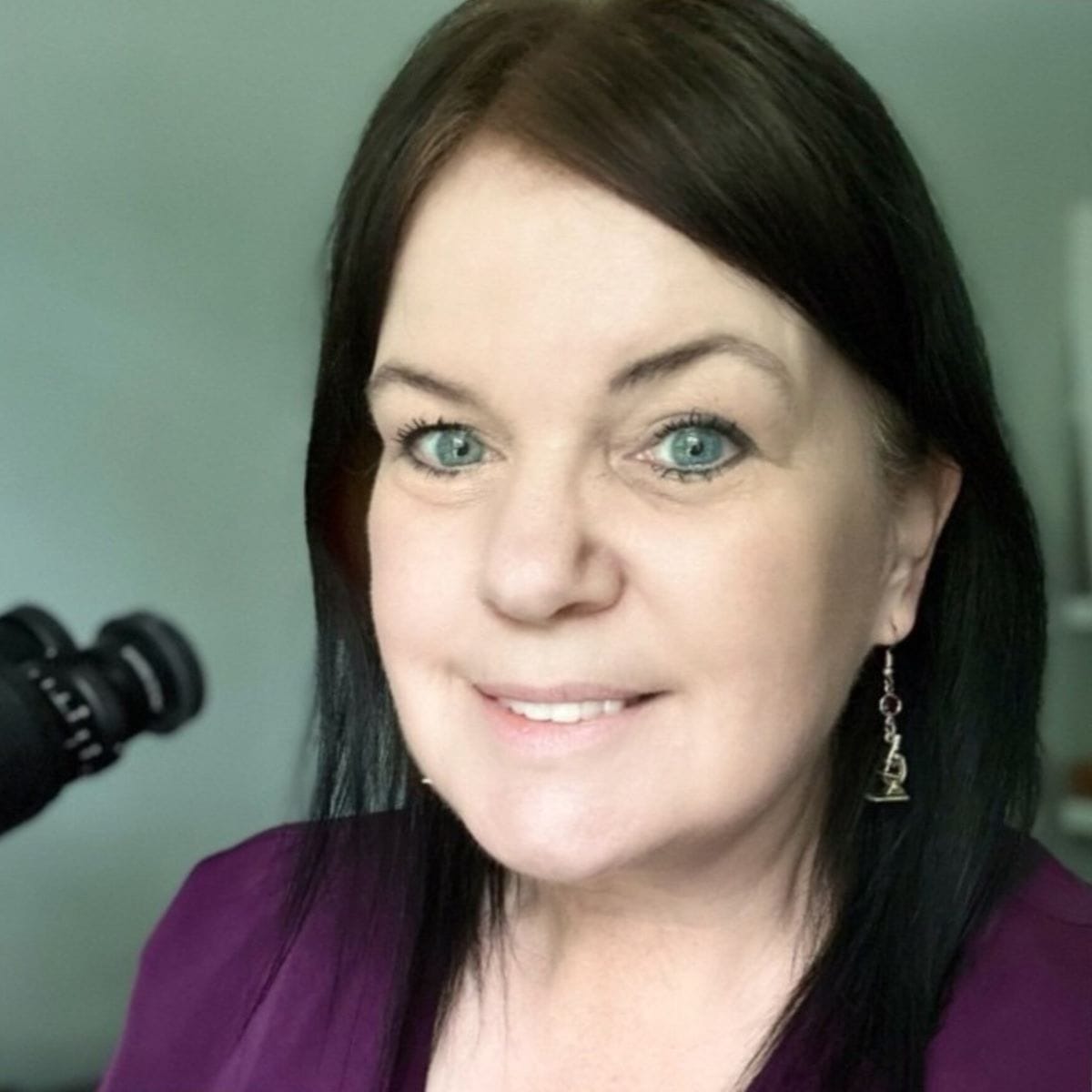 A woman with straight dark hair and blue eyes smiles at the camera. She is wearing a purple top and a single earring. A camera lens is visible in the background.