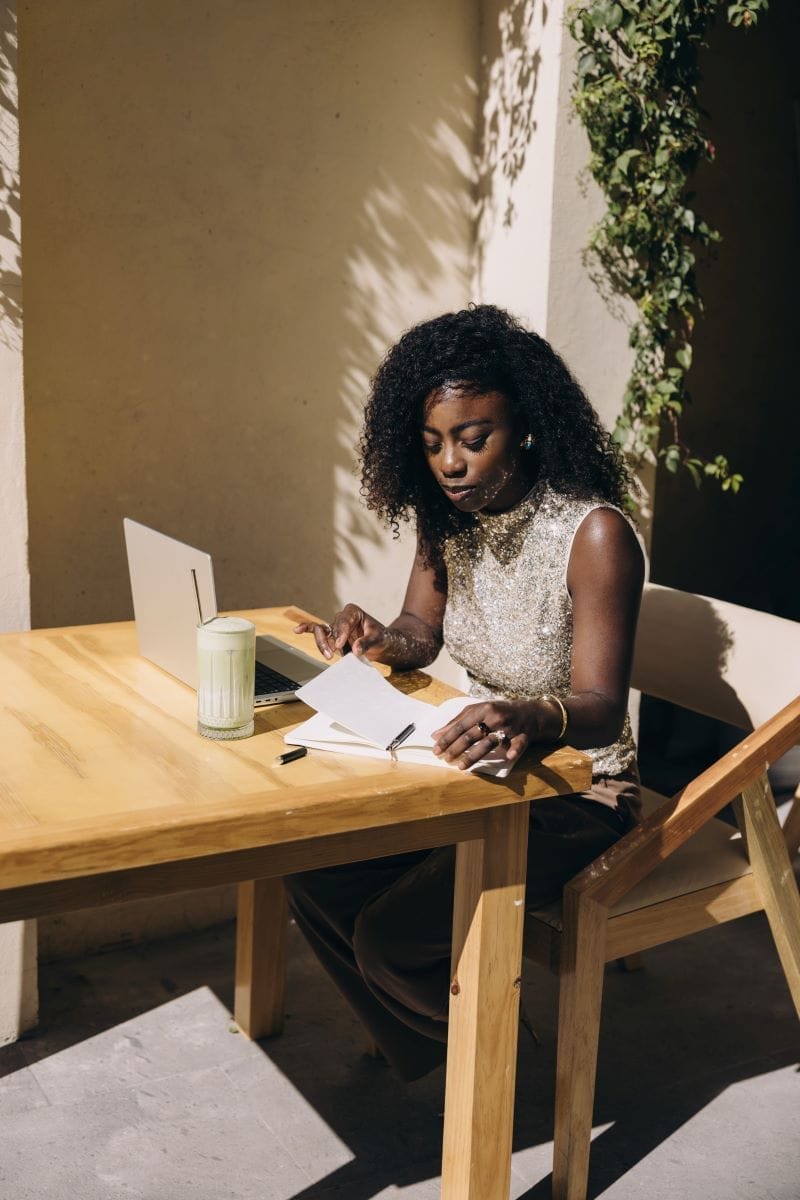 A woman sits at a wooden table outside, working with a laptop, an open notebook, and a drink, under sunlight near a leafy wall.