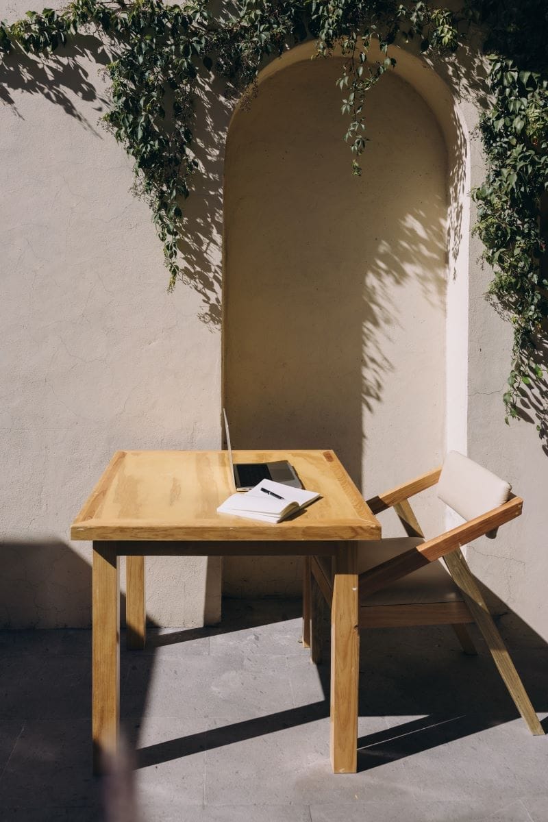 A wooden table and chair sit in sunlight against a beige wall with green vines; an open notebook and laptop are on the table.